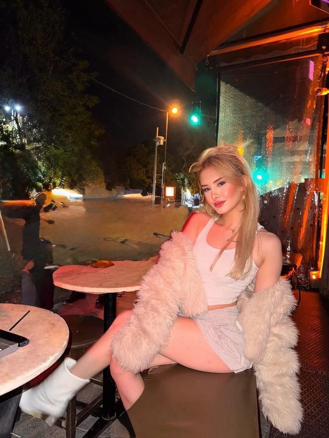 Young woman with blonde hair sits at an outdoor cafe table at night, wearing a white crop top, fluffy coat, skirt, and white boots under colorful city lights.",