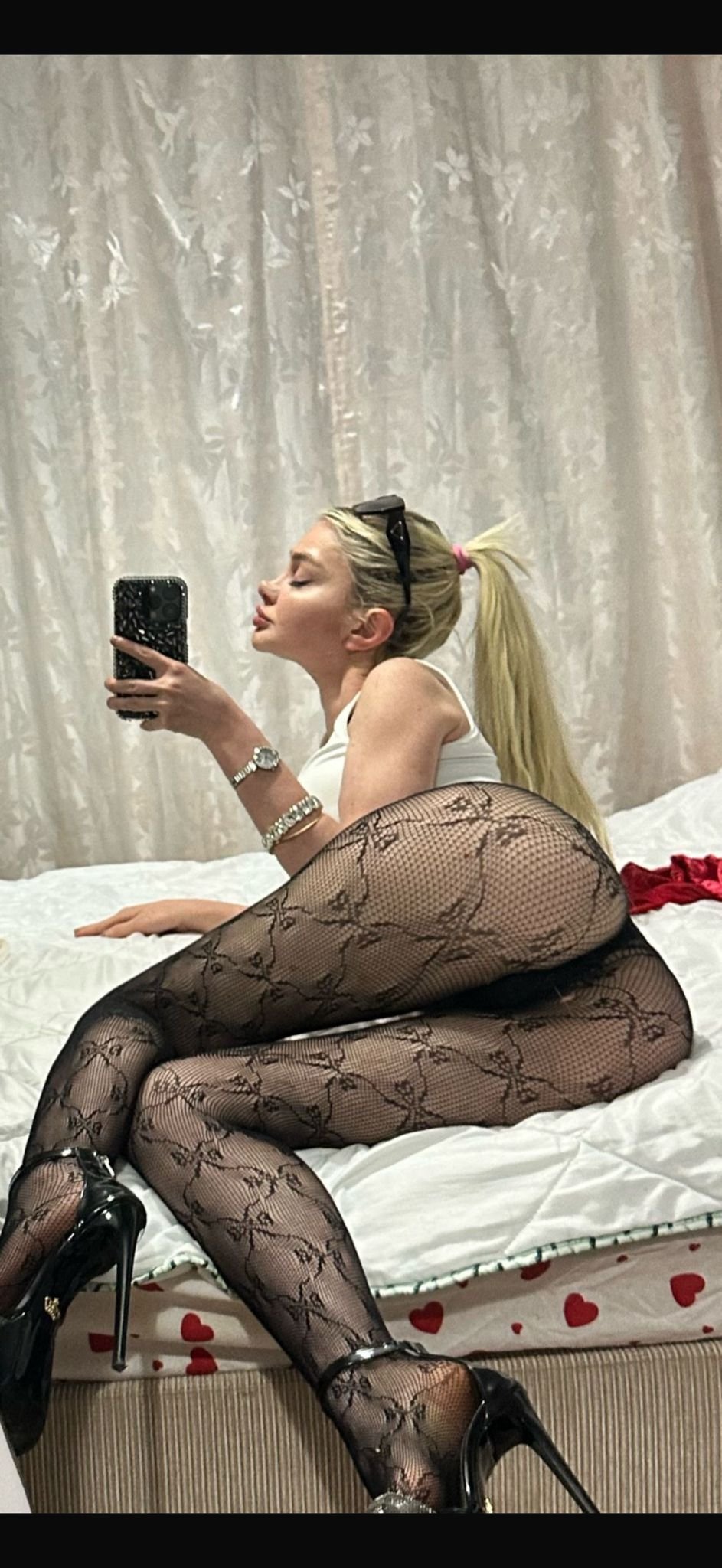 Side view of a woman on a bed taking a mirror selfie with her phone; wearing a white top, black floral-patterned fishnet tights, and black high heels, with a ponytail and sunglasses on her head.