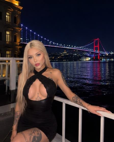 Woman with long blonde hair in a black cut‑out dress posing on a railing by a waterfront at night, with a lit suspension bridge in the background.