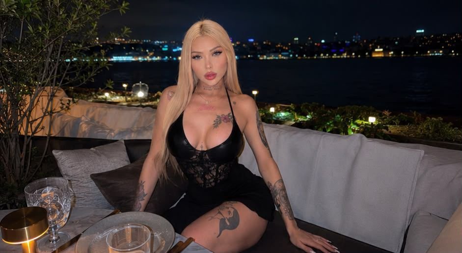 Blonde woman in a black lace outfit sits on an outdoor sofa at night, city lights in the background by a dining table with drinks.