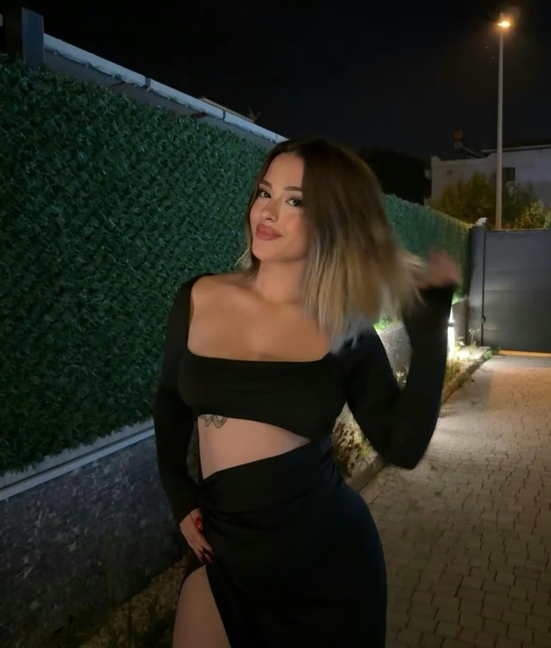 Woman in a black long-sleeve crop top and matching high-slit skirt posing at night beside a hedge. She smiles and flips her hair toward the camera.