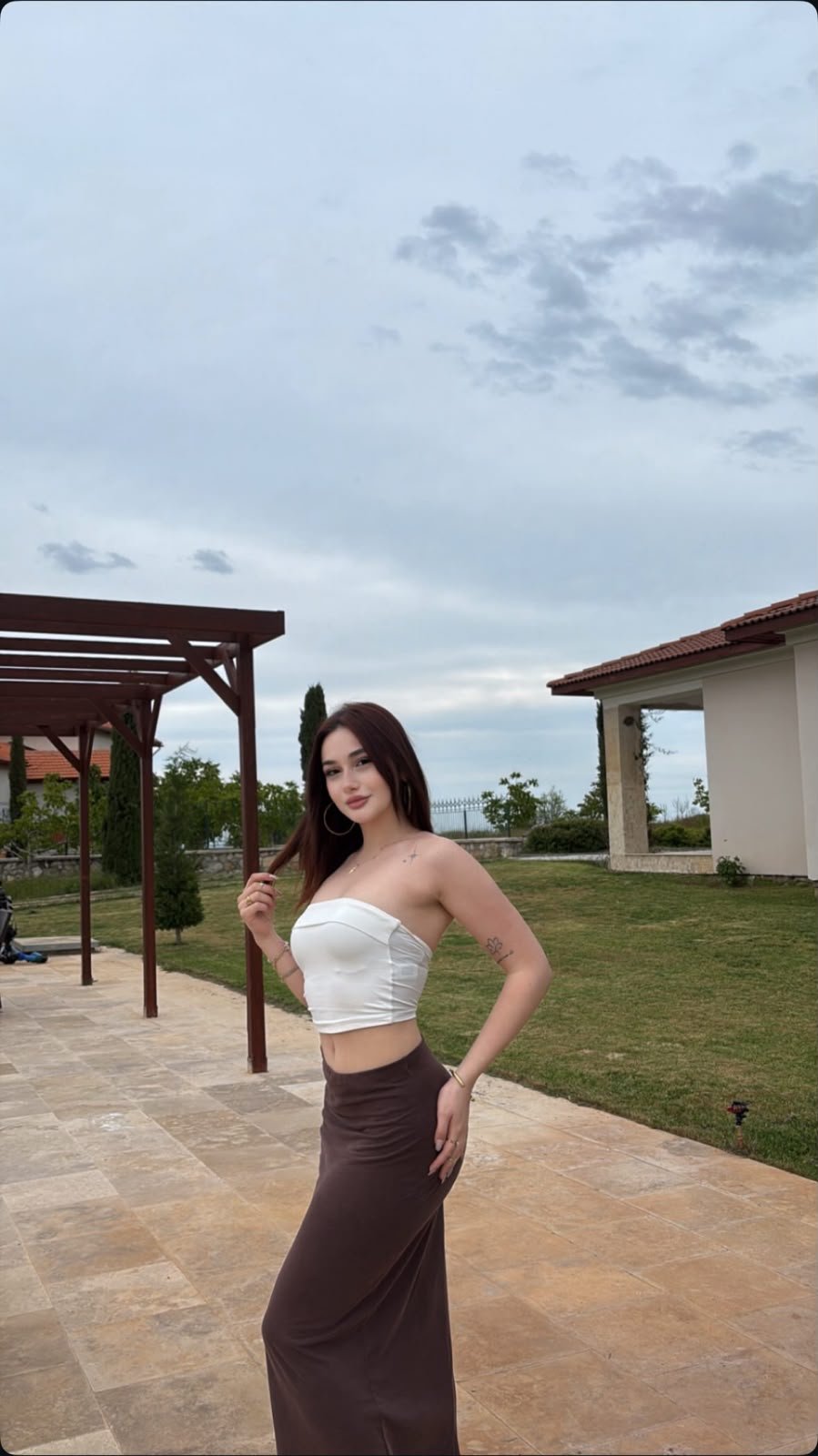 Woman posing outdoors in a white strapless top and brown maxi skirt on a stone patio, with a cloudy sky and pergola to the left.