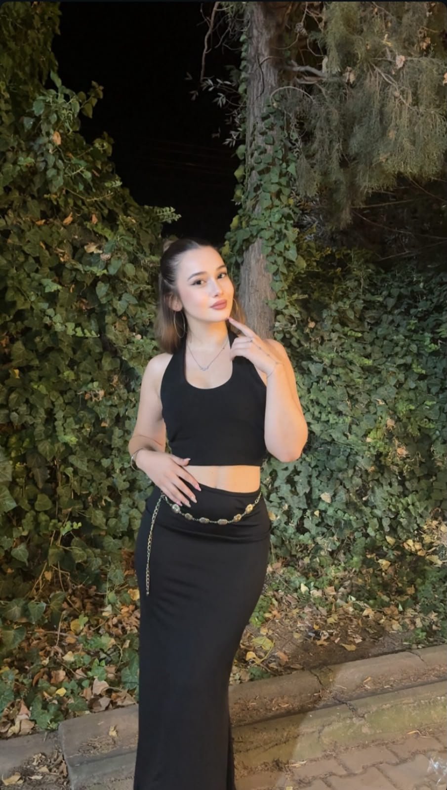 Woman in a black crop top and long black skirt posing outdoors at night beside an ivy-covered wall.