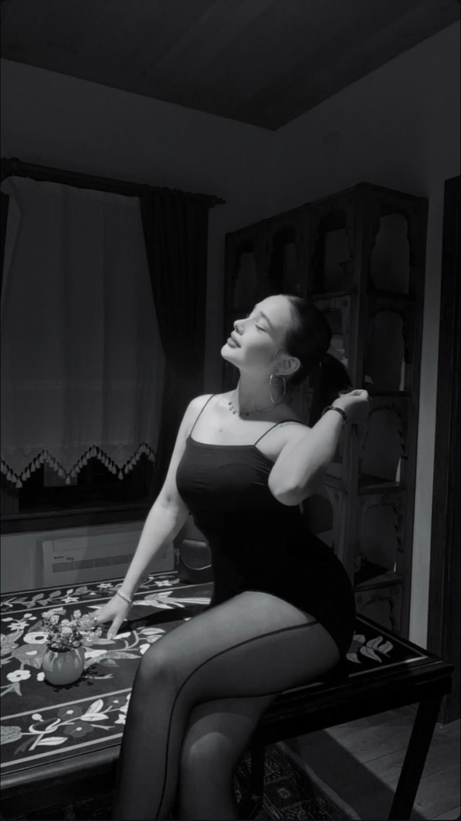 Woman in a black sleeveless dress sits on a table indoors, head tilted back and smiling.