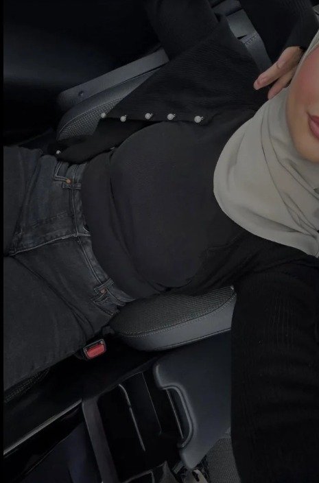 Person wearing a light gray hijab and black top sits in a car seat, with a red seat belt buckle visible in the foreground.