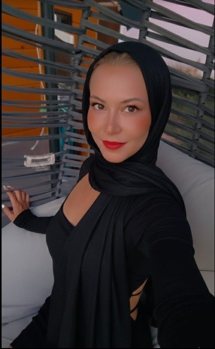Woman in a black hijab and outfit seated on a white couch, smiling at the camera with red lipstick.