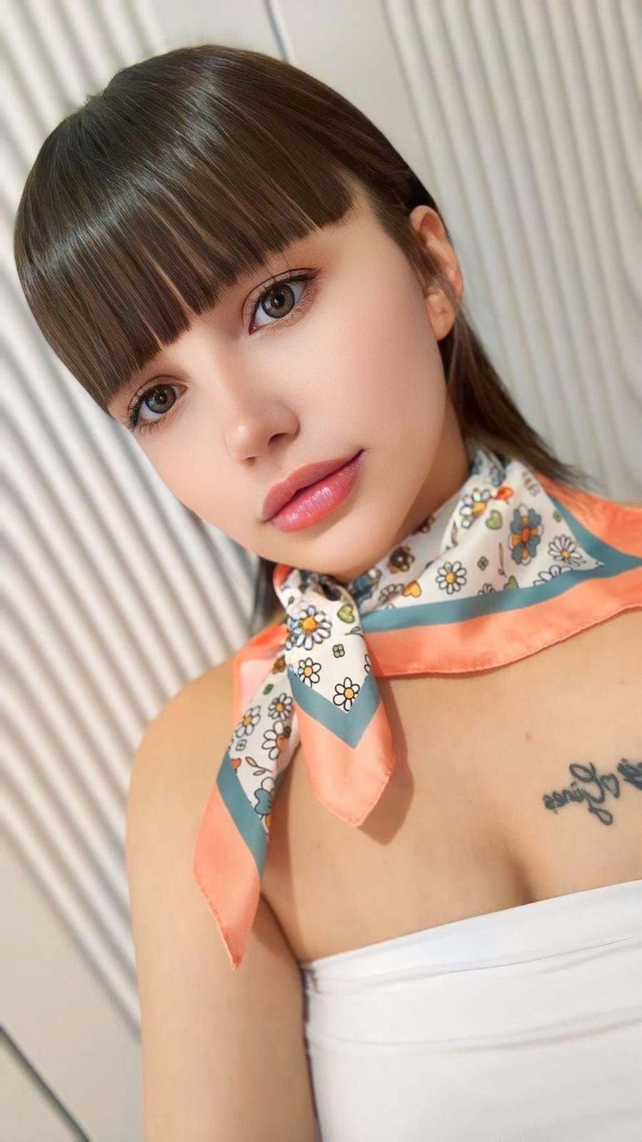 Young woman with brown hair and blunt bangs looks at the camera, wearing a floral scarf tied at the neck and a white strapless top.