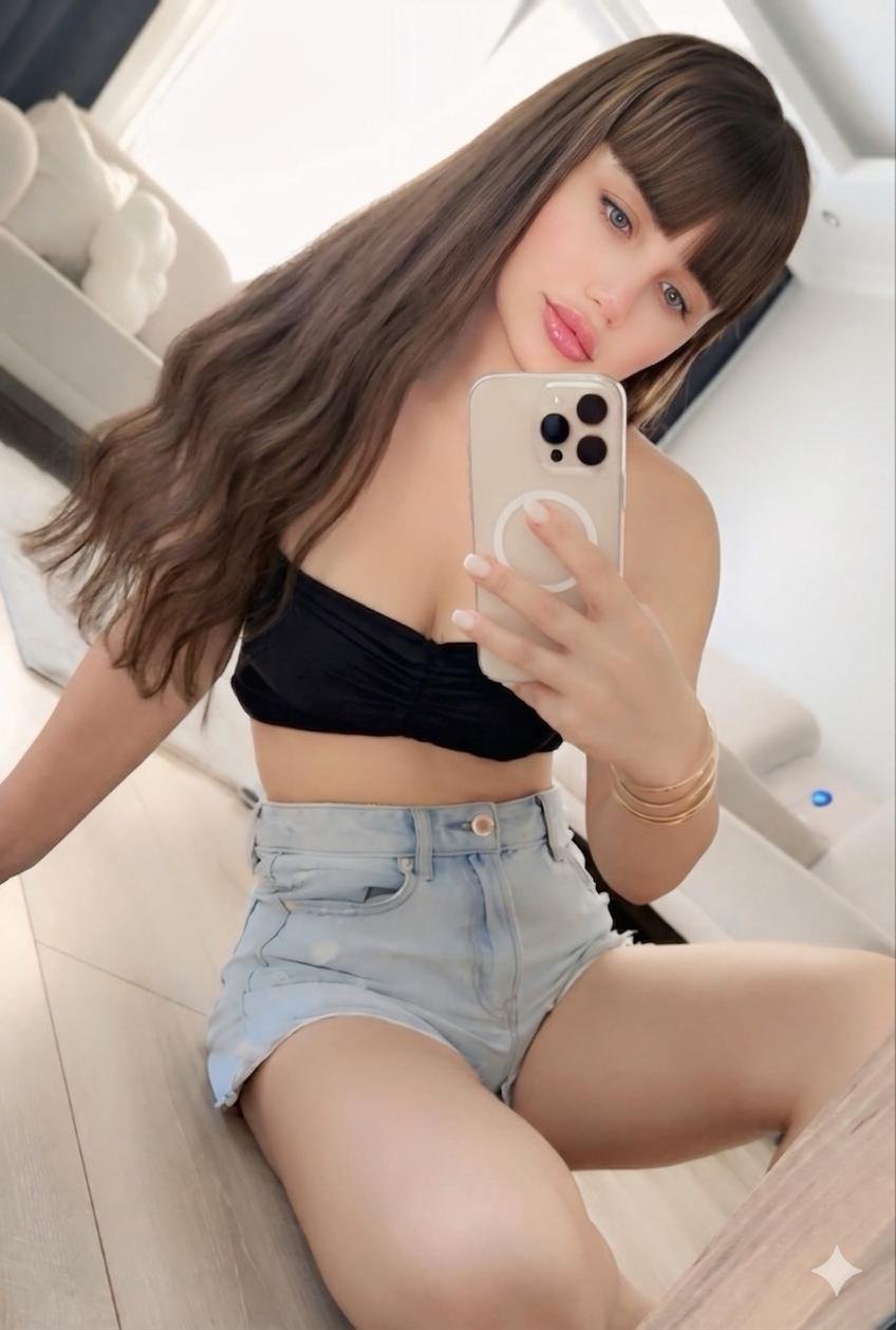 Young woman taking a mirror selfie in a bright room, wearing a black crop top and light denim shorts.