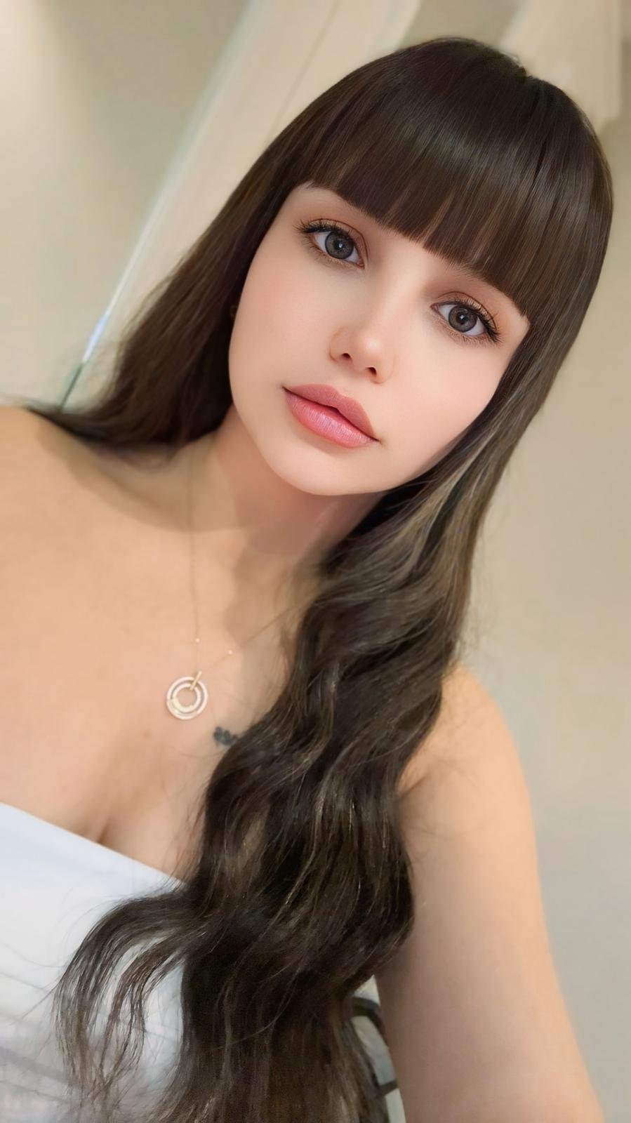 Portrait of a young woman with straight bangs, long dark wavy hair, wearing a circular pendant necklace and a white top.