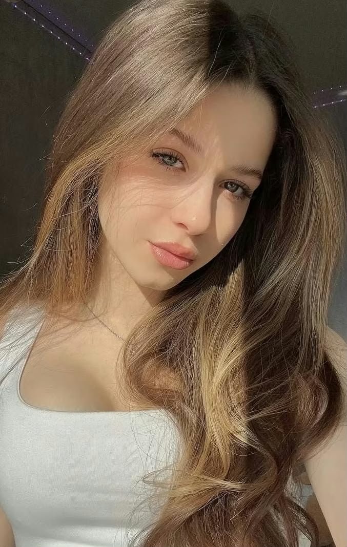 Young woman with long, light brown hair and subtle makeup gazing softly at the camera in sunlight, wearing a white tank top and a delicate necklace.
