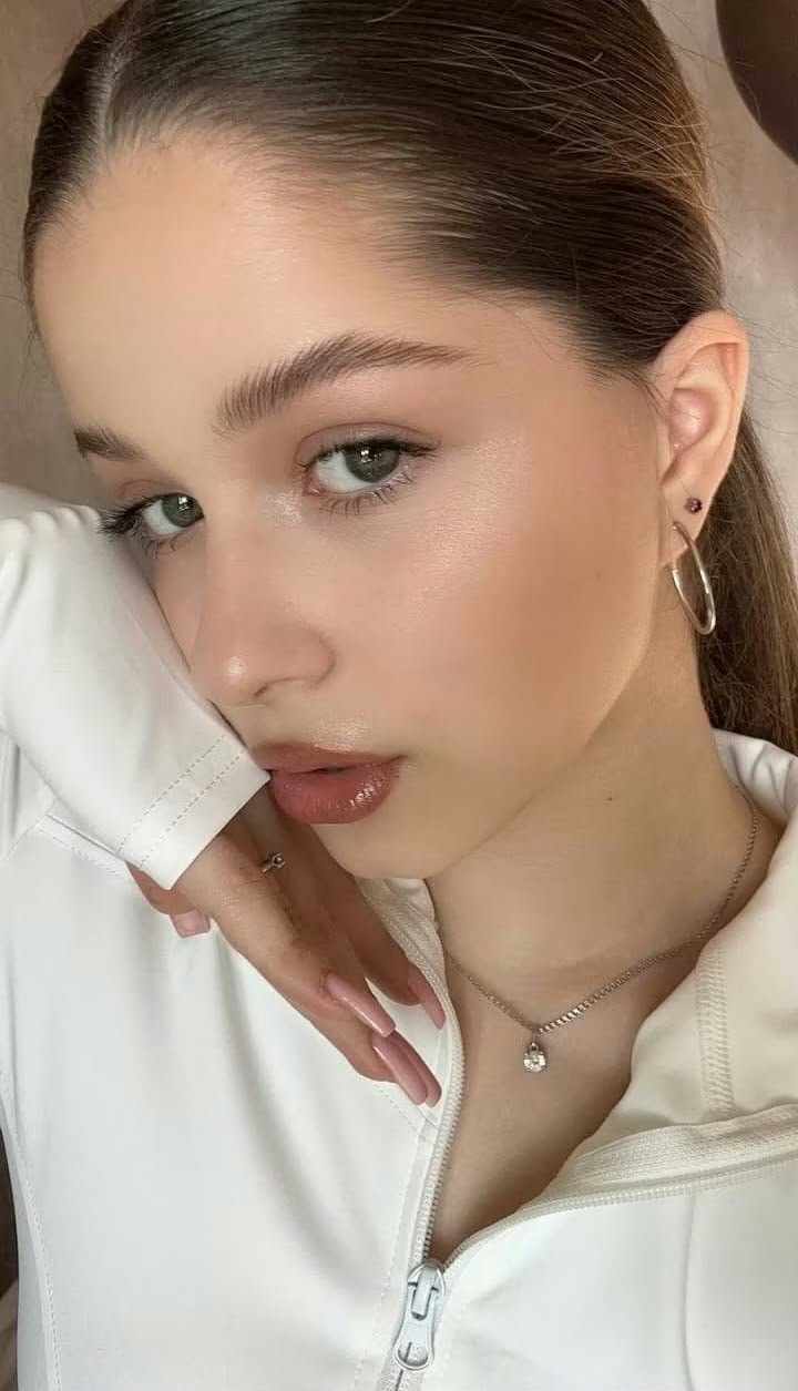 Close-up of a young woman with slicked-back hair, wearing a white zip jacket, hoop earring, and a pendant necklace, looking at the camera.