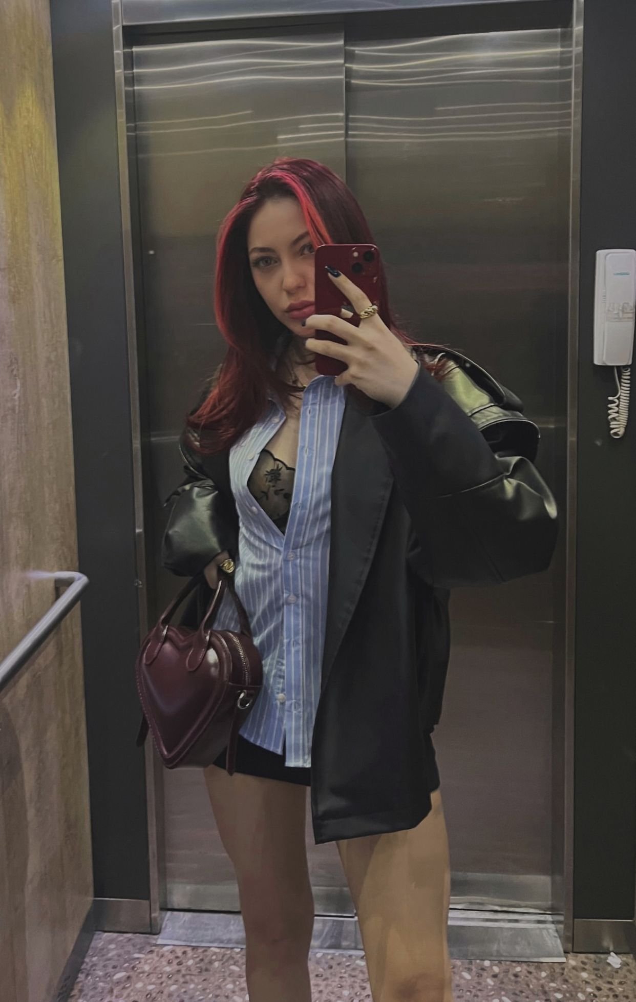 Woman with red hair taking a mirror selfie in an elevator, wearing a blue striped shirt under a black coat and carrying a maroon handbag.