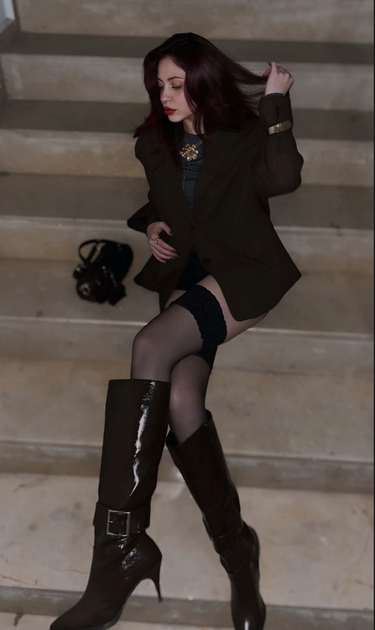 Woman with dark hair and red lipstick poses on a stairway in a brown blazer, dark top, sheer stockings, and tall brown boots; a black handbag sits on the step behind her.