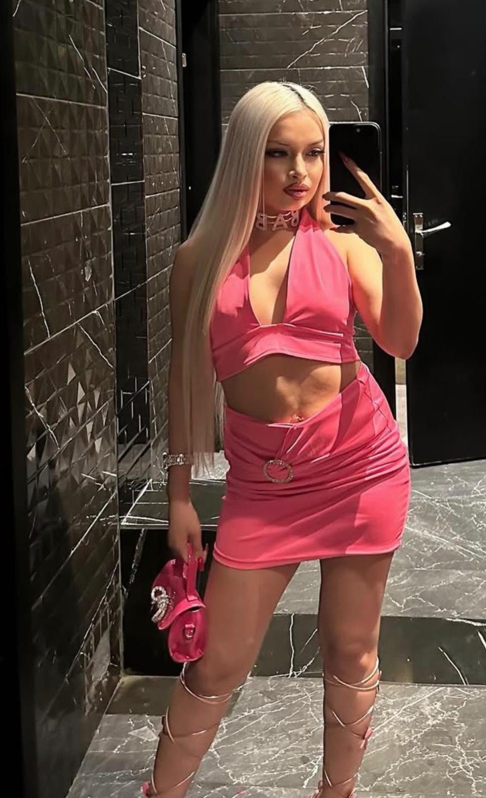 Person taking a mirror selfie in a marble-tiled bathroom, wearing a pink halter crop top and matching mini skirt with long blonde hair.