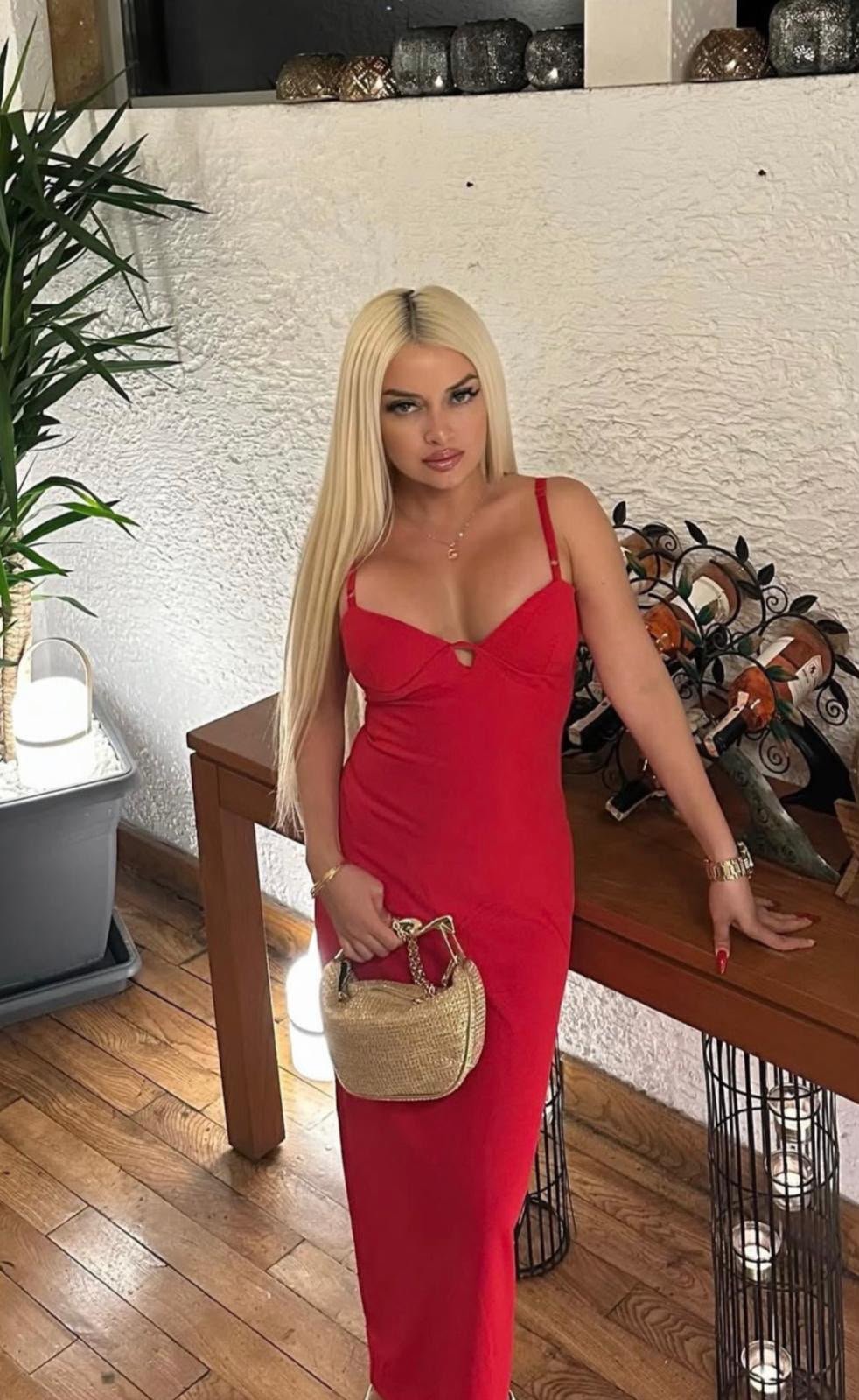 Woman with long blonde hair wearing a red spaghetti-strap dress posing indoors beside a wooden table, holding a small woven straw handbag.