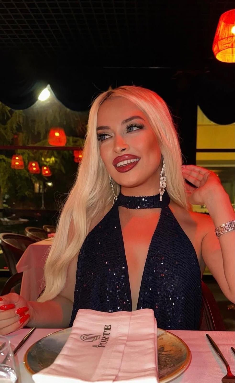 Woman with long blonde hair in a sparkly navy halter dress at a candlelit restaurant, smiling.