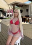 Woman with long blonde hair in a red bikini under a white mesh cover, posing on a chair at an outdoor venue with a stage in the background.