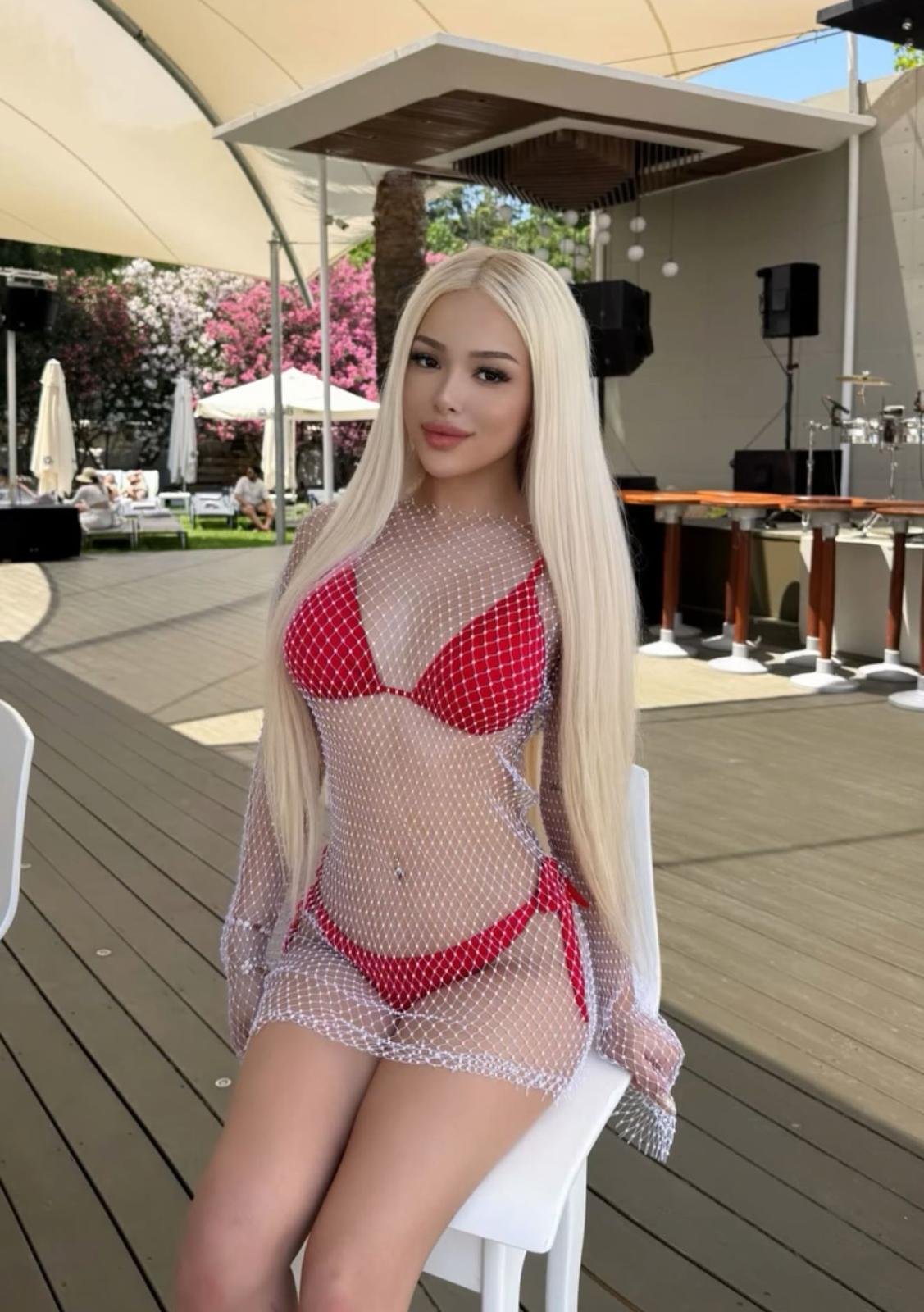 Woman with long blonde hair in a red bikini under a white mesh cover, posing on a chair at an outdoor venue with a stage in the background.
