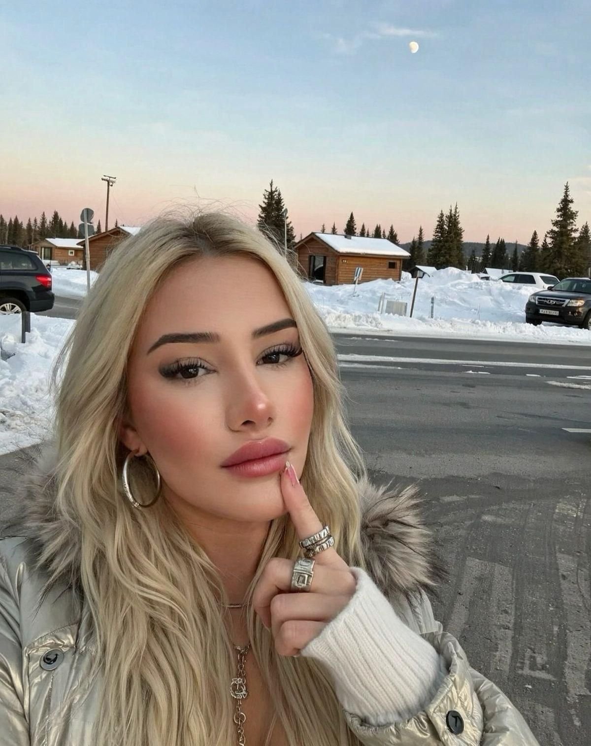 Close-up selfie of a blonde woman in a winter coat with fur trim, posing with a hand near her chin in a snowy parking area.