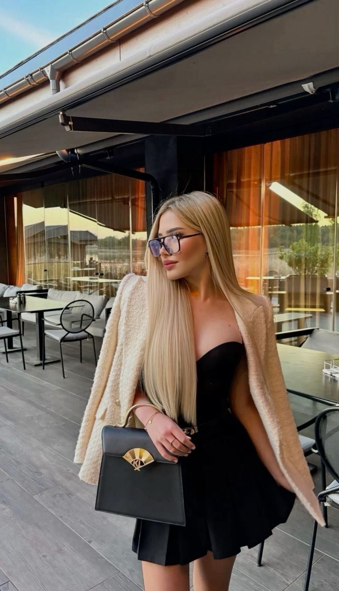 Stylish woman with long blonde hair in a black dress and beige coat, wearing sunglasses, holding a black handbag at an outdoor cafe.