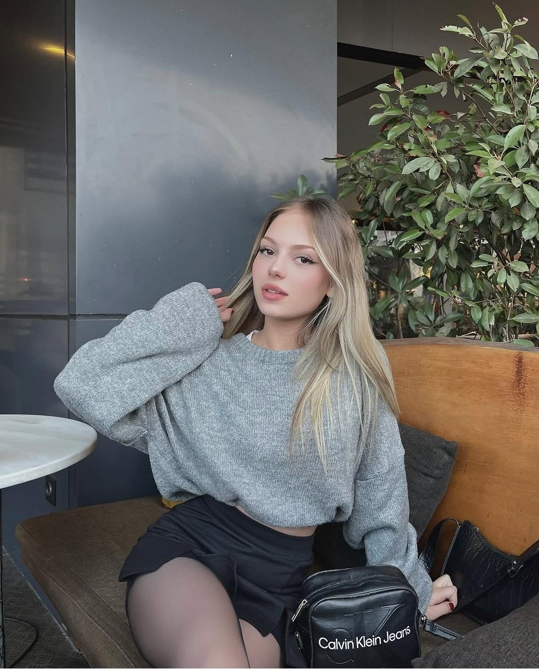 Young woman with blonde hair wearing a gray knit sweater and black shorts sits on a wooden bench at an outdoor cafe, with a black Calvin Klein bag nearby.
