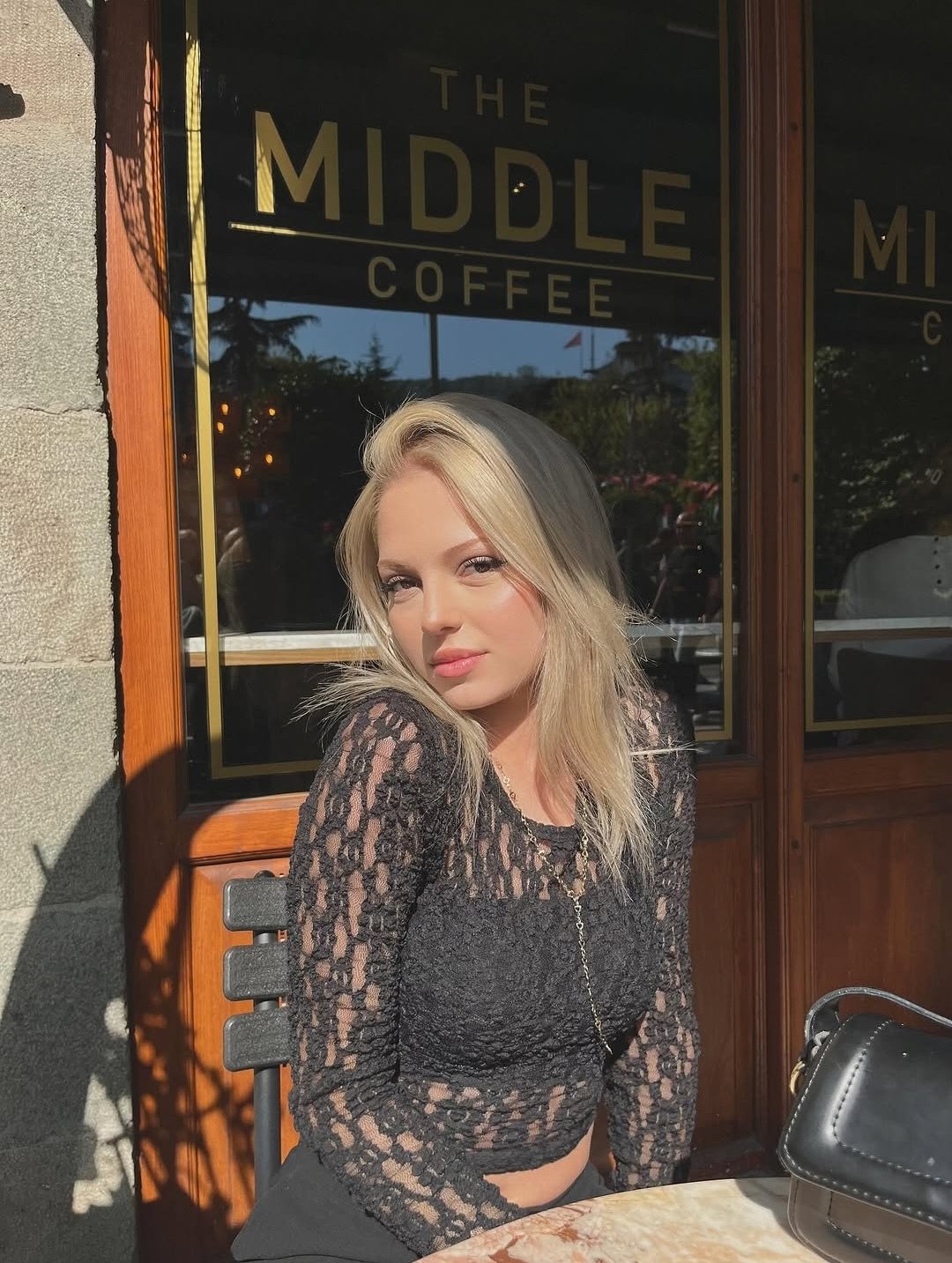 Blonde woman in a black lace top sits at an outdoor café table in sunlight, with 'THE MIDDLE COFFEE' sign visible behind her.