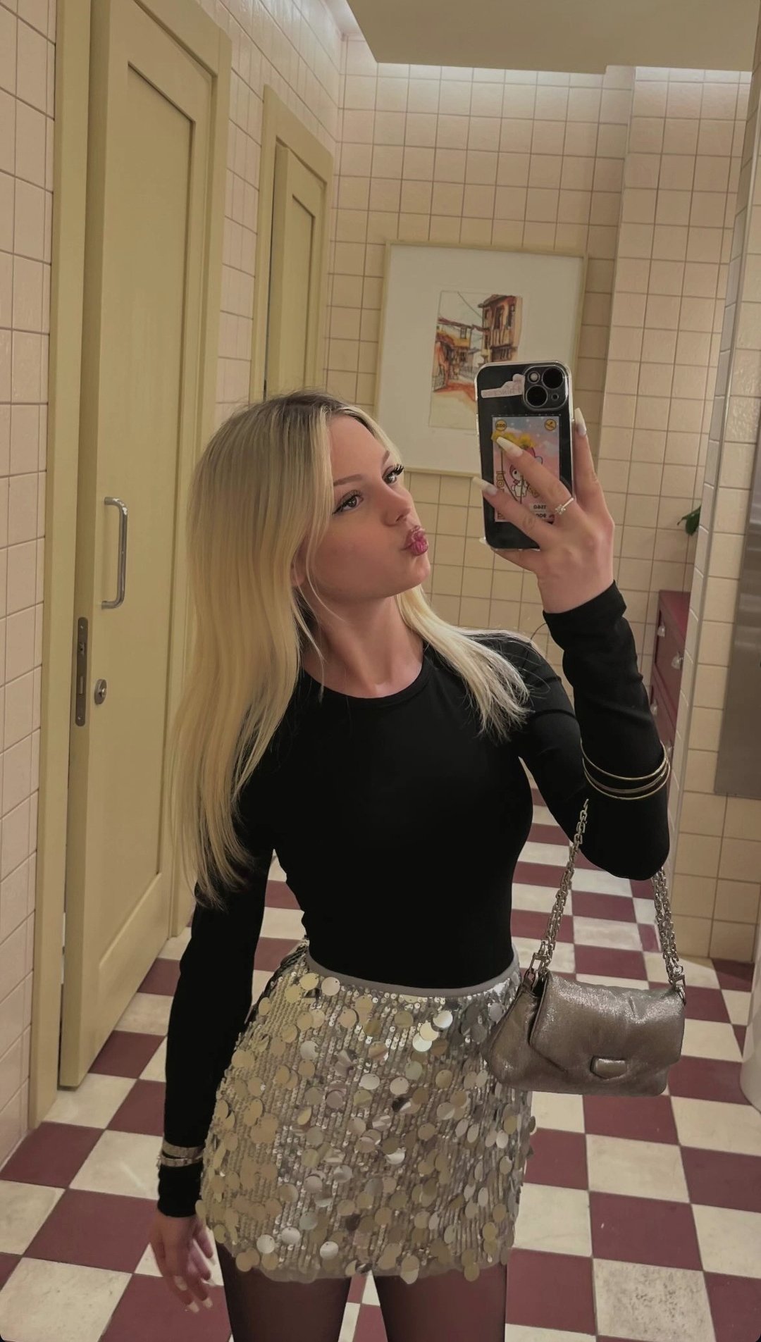 Woman with blonde hair in a black long-sleeve top and silver sequin skirt takes a mirror selfie in a tiled restroom, holding a purse on her shoulder.