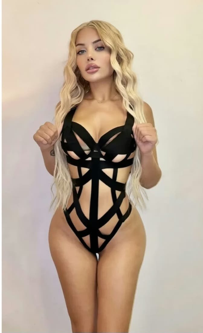 Woman with long wavy blonde hair wearing a black strappy one-piece swimsuit, facing the camera against a light background in a confident pose.