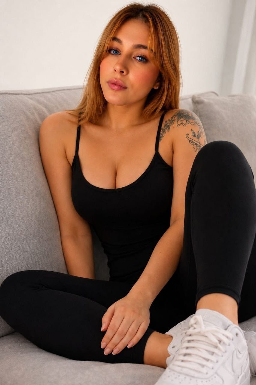 Young woman with light brown hair sits on a gray couch, wearing a black tank top, black leggings, and white sneakers; tattoo on her left shoulder.