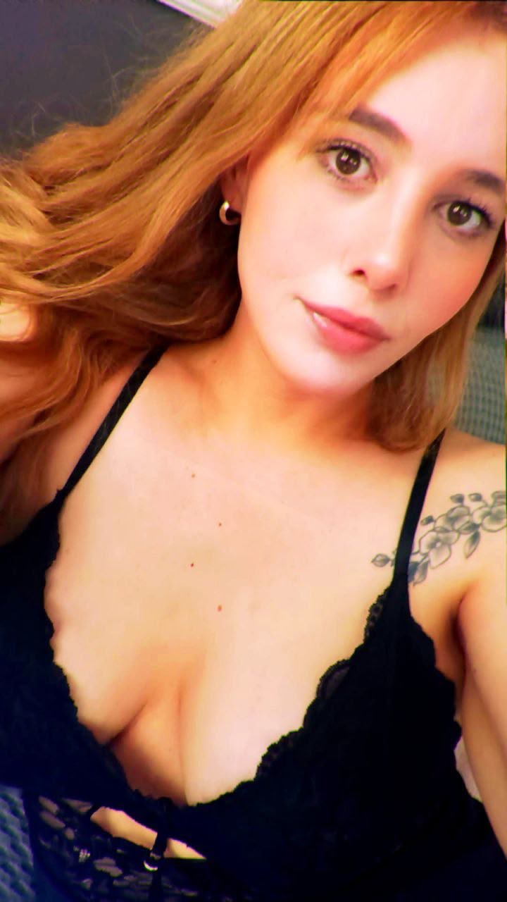 Close-up selfie of a woman with orange hair wearing a black lace top, showing a tattoo on her left shoulder.