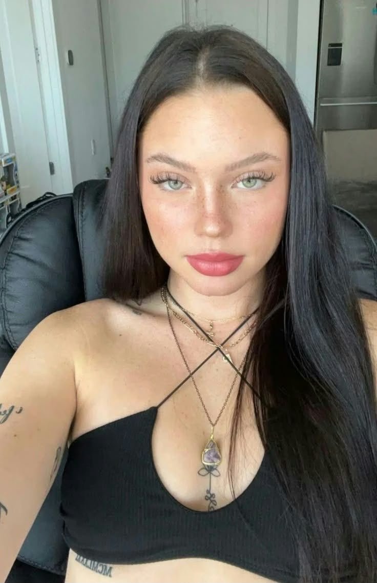 Selfie of a young woman with long dark hair wearing a black halter top, layered necklaces, and visible tattoos on her arms.