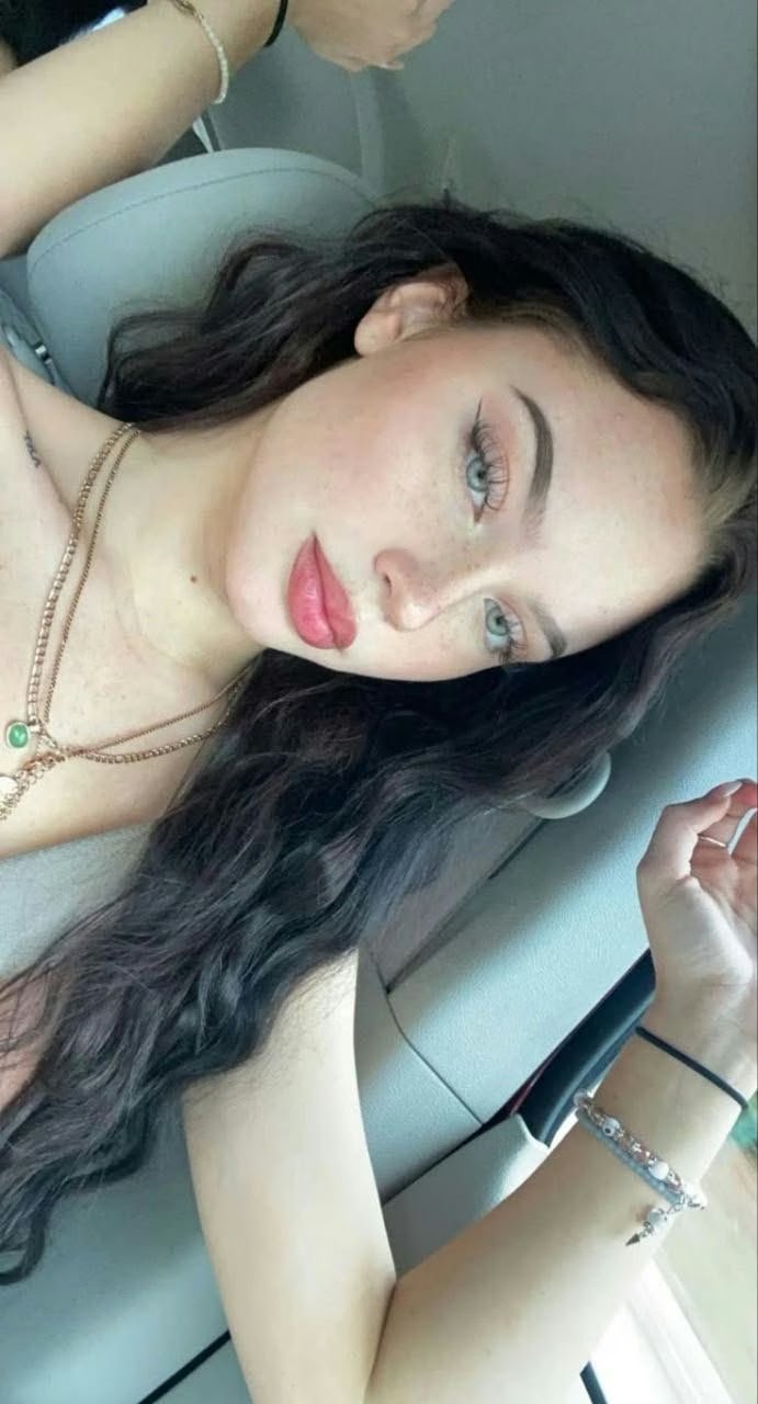 Woman with long dark hair in a car, wearing pink lipstick and layered necklaces, looking at the camera.
