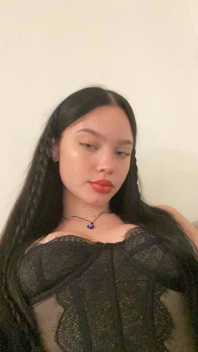 Portrait of a young woman with long black hair and braided sides, wearing a black lace top and a small blue pendant necklace against a pale wall.