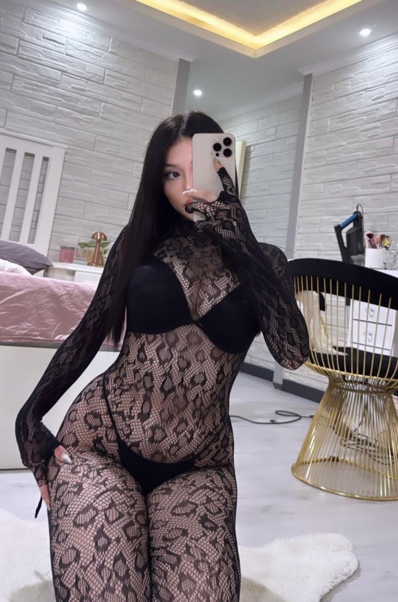 Woman in a black lace bodysuit posing for a mirror selfie in a bright, modern bedroom setting.